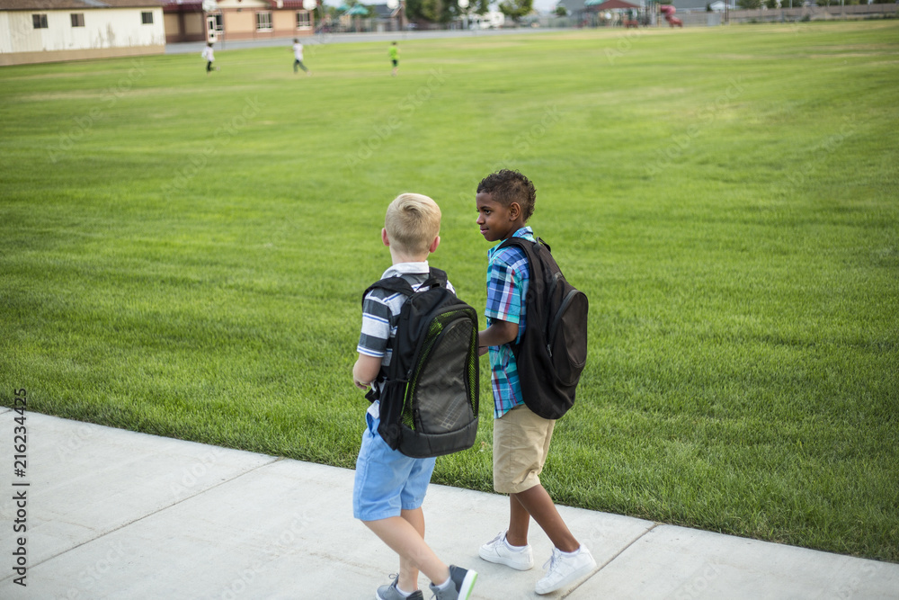 Two diverse school kids walking and talking together on the way to school. Back to school photo of diverse school children wearing backpacks in the school yard. Safety tips for walking home from school.