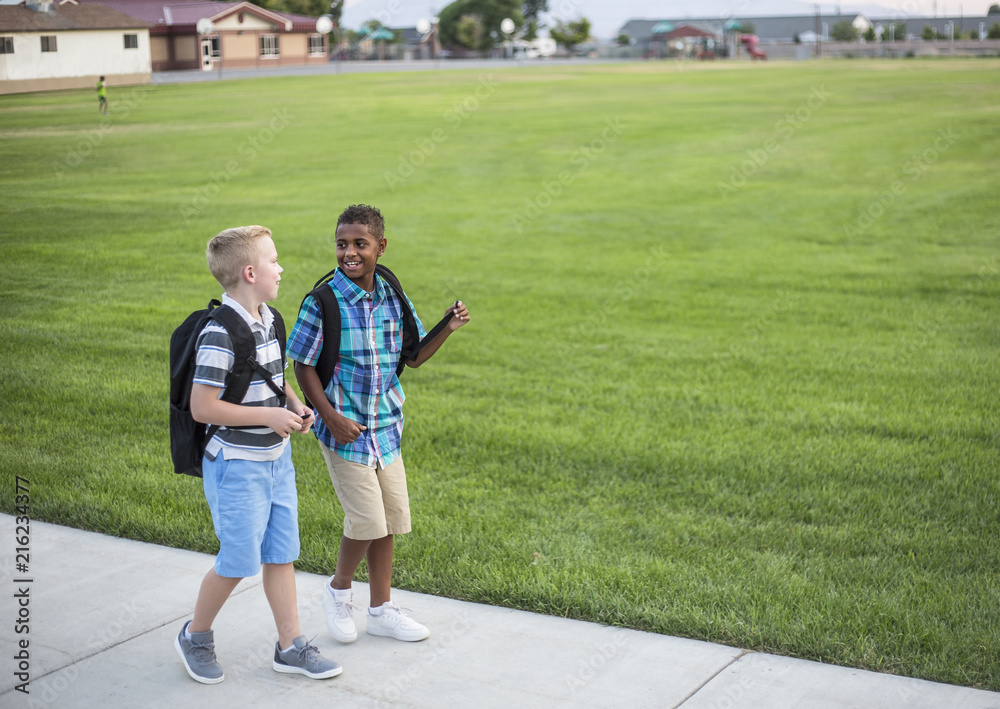 Two diverse school kids walking home together after school and talking together. Back to school photo of diverse school children wearing backpacks in the school yard