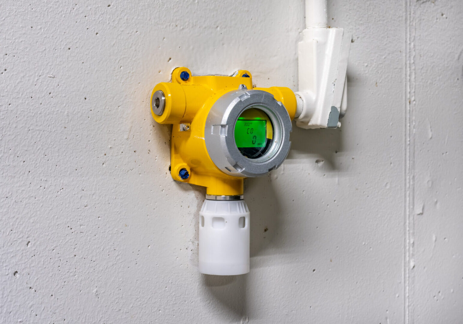 Yellow gas detector sensor mounted on a wall with white piping.